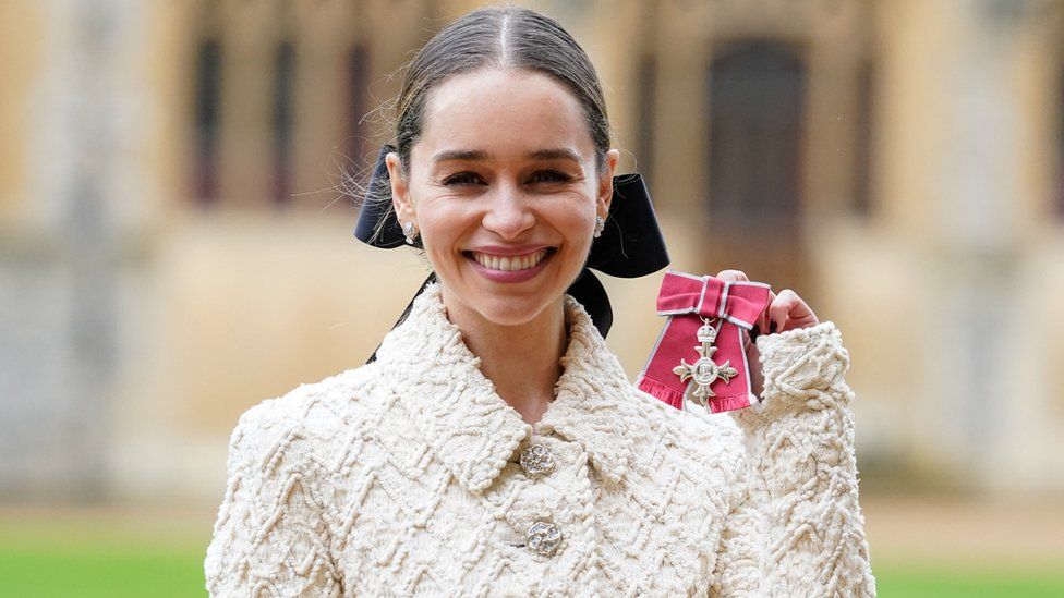 Emilia Clarke smiles while holding her MBE