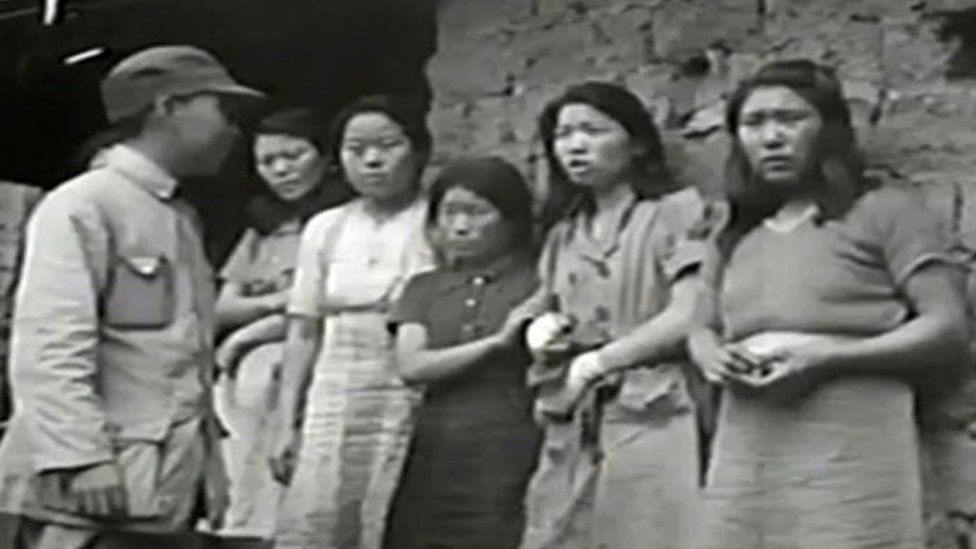 Korea women and girls lined up against a wall with a Japanese soldier looking at them.