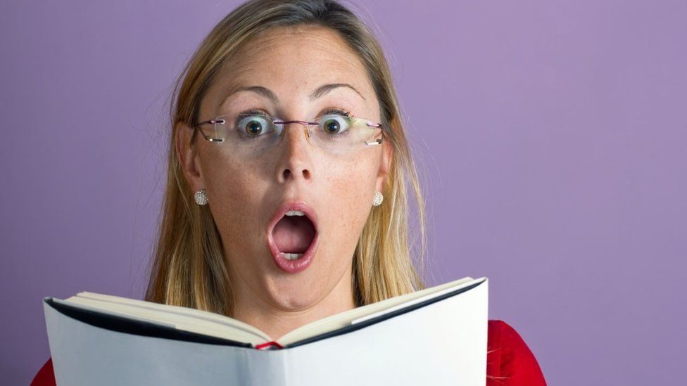 photo shows a woman reading a book with a shocked expression