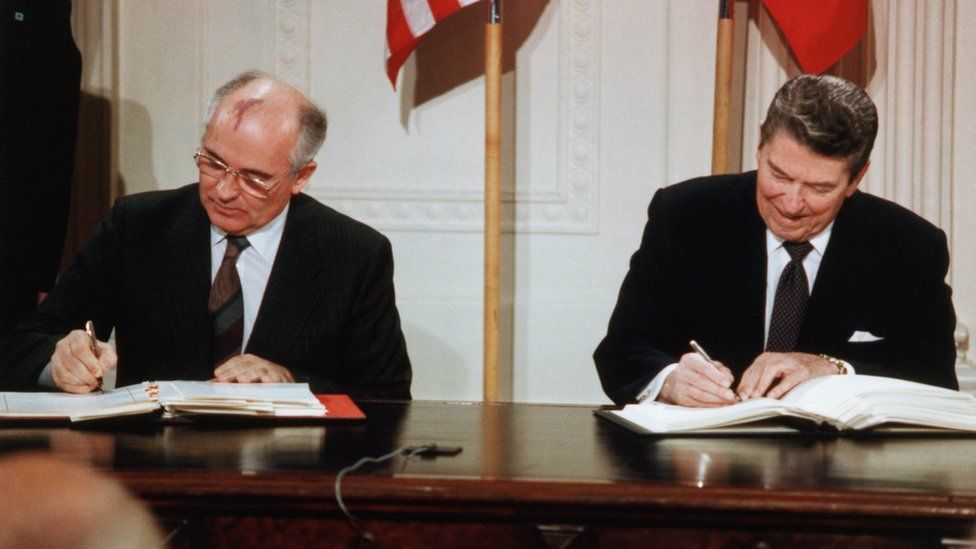 Reagan and Gorbachev Signing Arms Limitation Agreement at a table in front of their nations' flags.