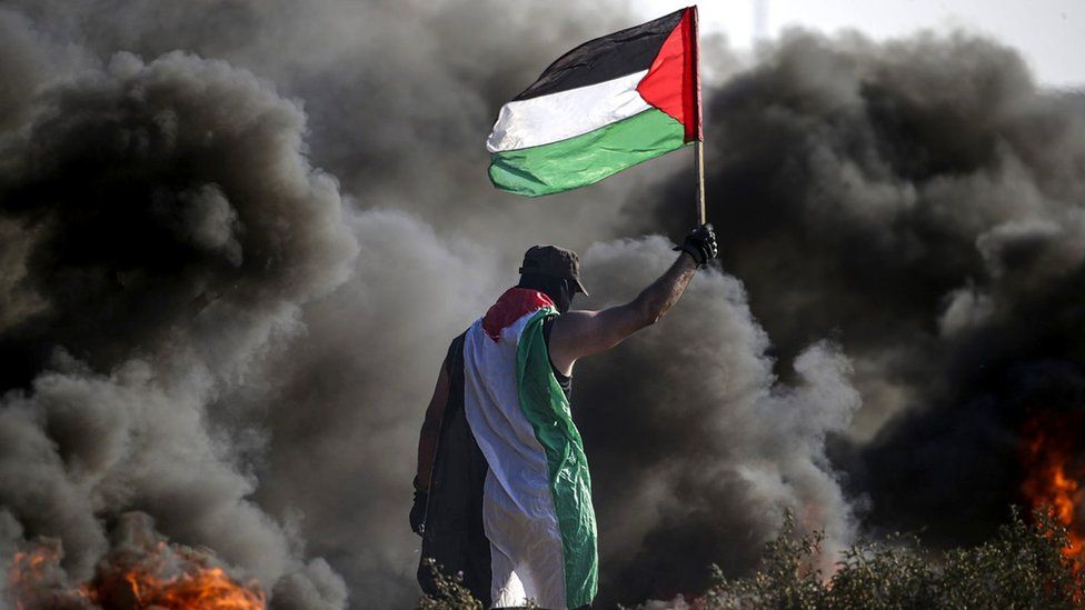 Палестинский протестующий машет палестинским флагом, стоя возле черного дыма, поднимающегося от горящих предметов, во время акции протеста возле ограждения по периметру сектора Газа-Израиль (22 сентября 2023 г.)