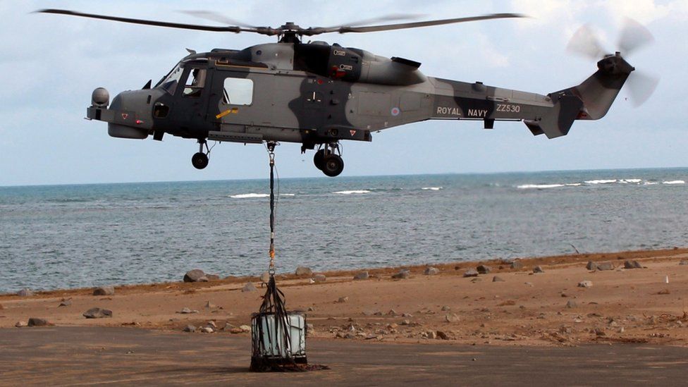 A helicopter hovers above a runway as aid is lowered down