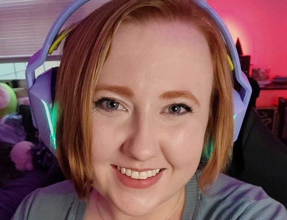 Ashlyn is smiling, wearing light purple gaming headphones which emanate a green light from the ear cups. The headset covers her reddish hair, which is about chin-length. She's in a room which appears to have gaming memorabilia just visible in the background. One corner is bathed in a soft red light.