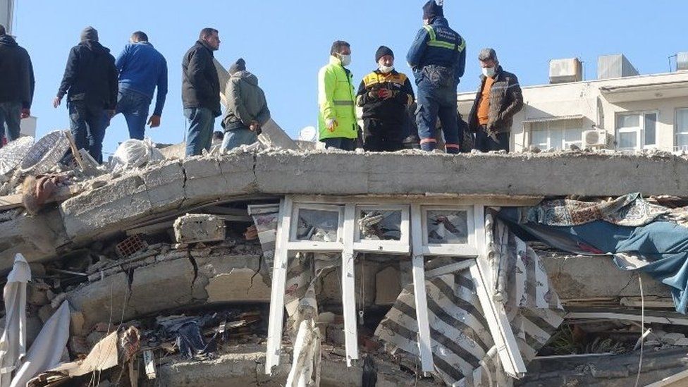 People stand on top of rubble. An intact window is visible in the layers of rubble.
