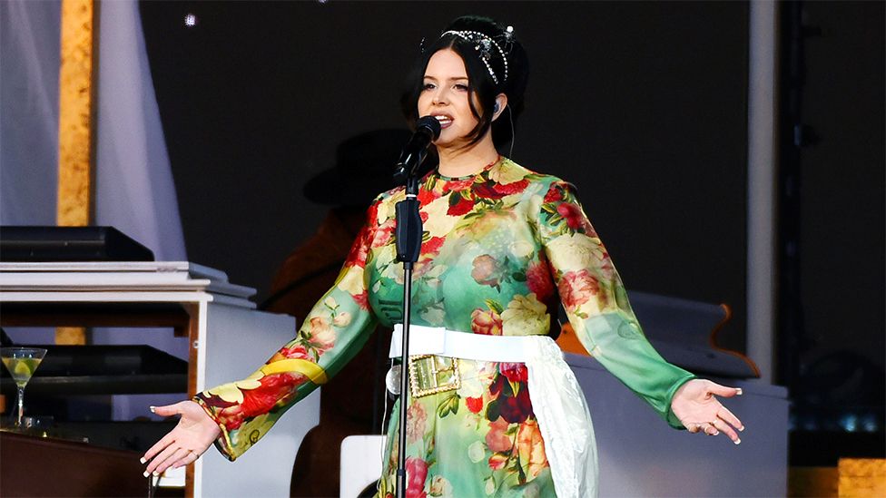 Lana Del Rey singing on stage, she is wearing a green floaty dress and her arms are open. She is wearing her black hair up.