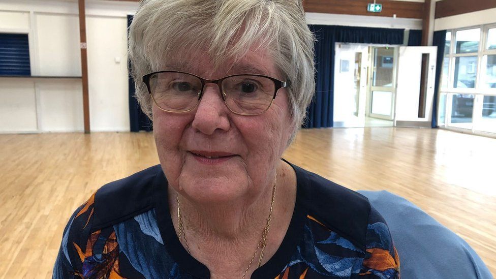 A lady in her 80s is looking at the camera. She is wearing glasses and a black top with orange and blue flowers. She is sat in a hall where she has just done a Tai Chi session.