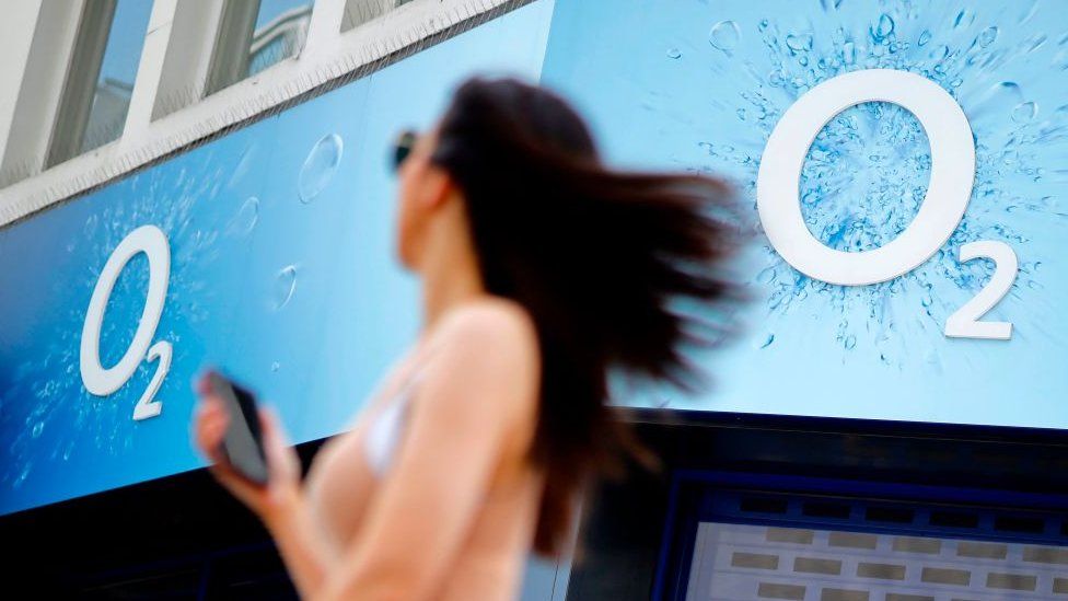 A woman holds a mobile phone as she walks past an O2 mobile phone store