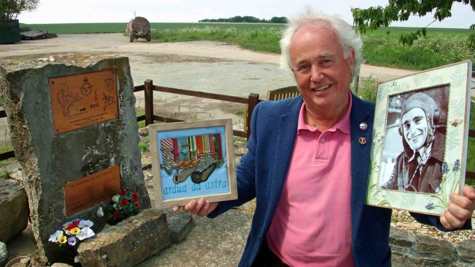 Robert Seymour standing next to the stone memorial at Tarrant Rushton and holding a framed black and white photo of his father in one hand and a frame containing his medals in the other hand