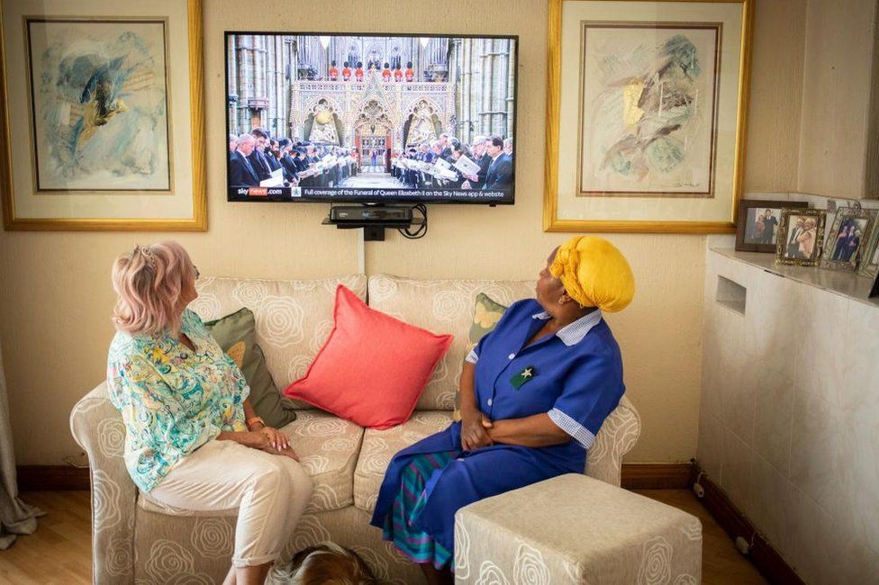 Two women sitting on a sofa in a home. Their heads are turned looking at the TV screen which shows the Queen's funeral procession. One woman is wearing a blue uniform with a yellow head wrap and the other is wearing trousers and a blue-ish shirt.
