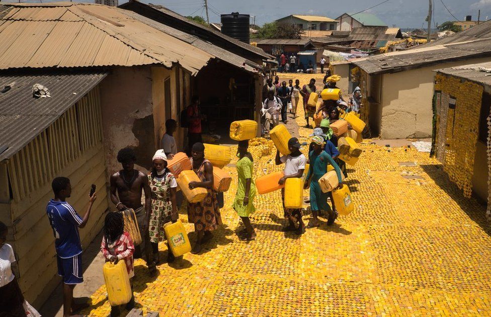 People queuing with jerrycans on yellow tapestry created by artist Serge Attukwei Clottey on a road in La - Accra, Ghana