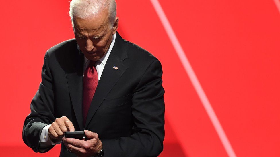 Joe Biden looks down at his smartphone while standing against a giant stage drop background in bright red