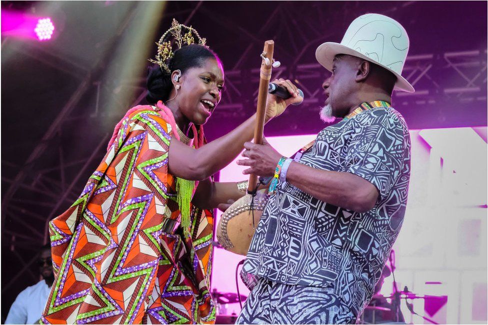 Woman singing in African attire with a crown on her head. Man is next to her in a hat with an instrument.