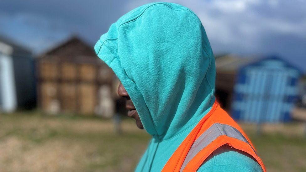 Offender in turquoise hoodie and fluorescent orange jacket by beach huts