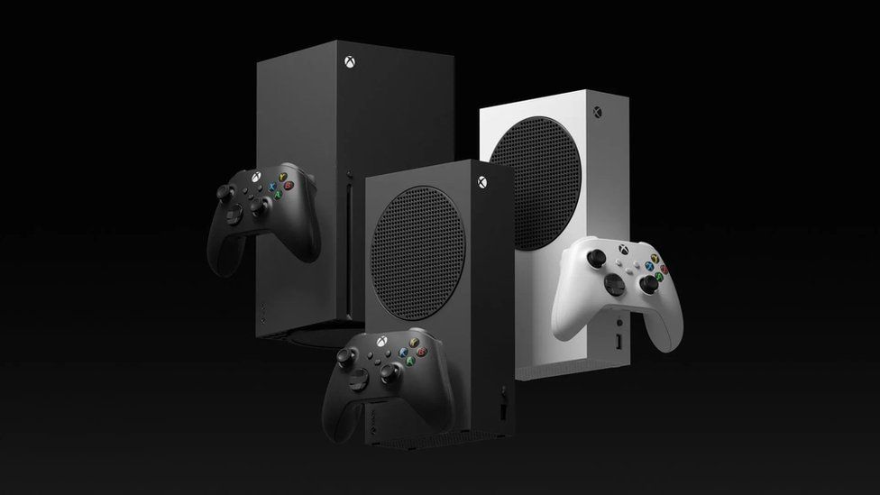 Three Xbox consoles - The Series X, the Series S (in white) and the new carbon black Series S are shown against a black background. They are arranged closely, each overlapping the other. Each has the corresponding controller pictured next to it - they appear to be floating next to the machines.