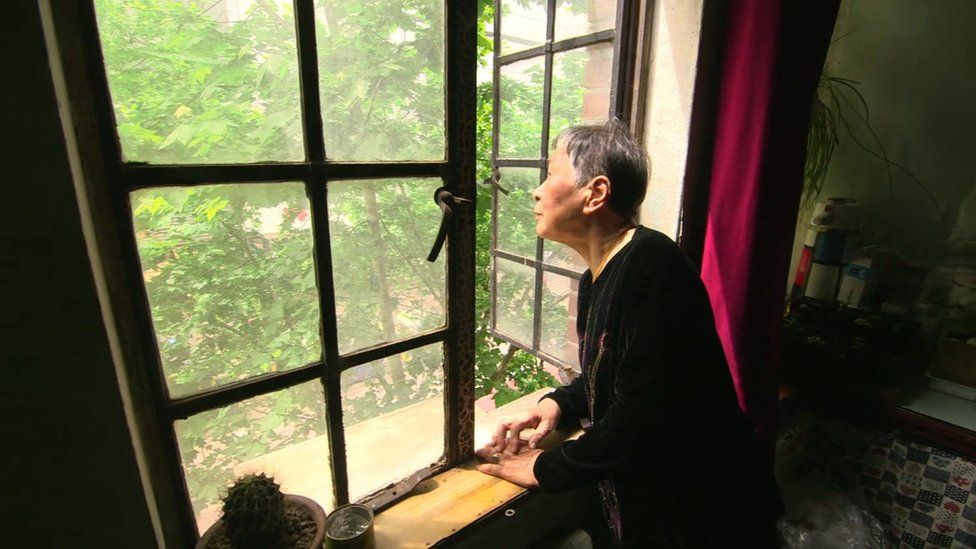 An older Chinese lady looks out from the window of her home