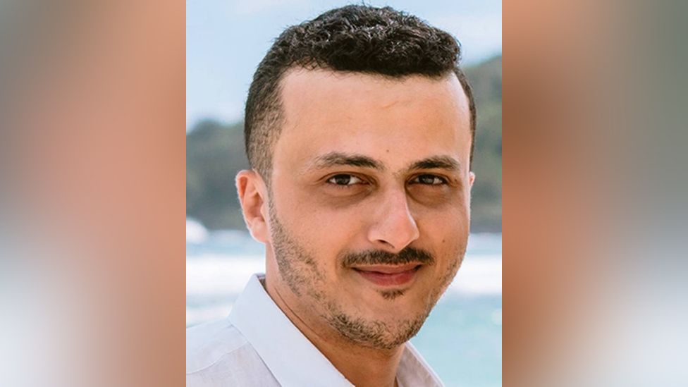 A headshot of Mohammed Al Imam in front of a beach/sea backdrop. He's got neatly trimmed, short black hair and a think, dark, moustache. He's wearing a white shirt with an open collar, and smiling slightly.