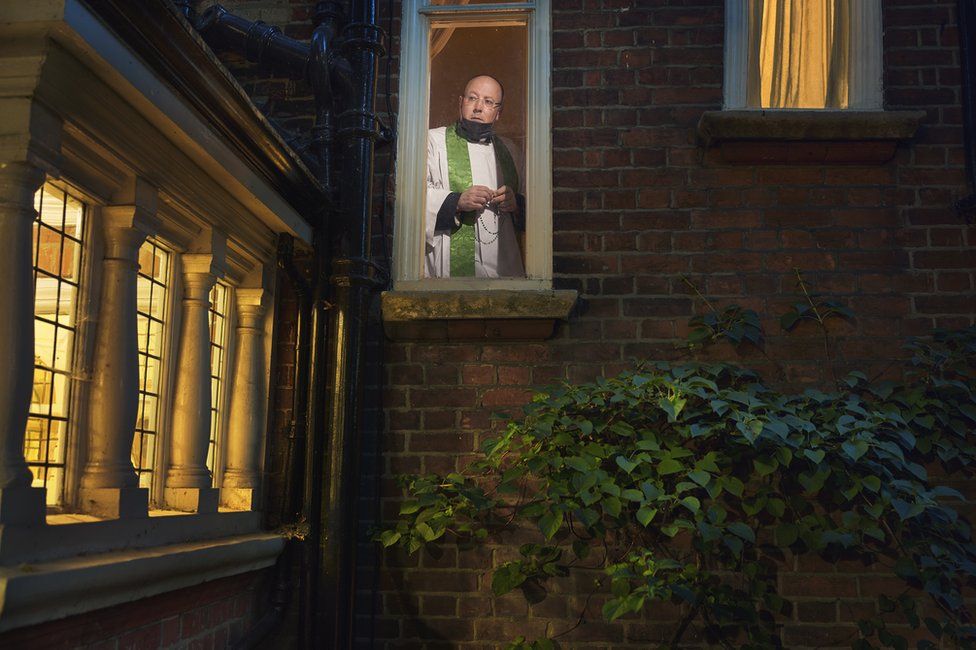 A priest stands looking out the window of a house in London in a stylised portrait