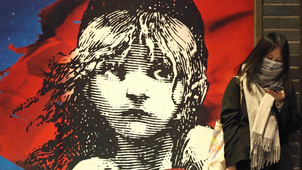 Женщина в маске стоит перед плакатом Les Miserables в лондонском Вест-Энде