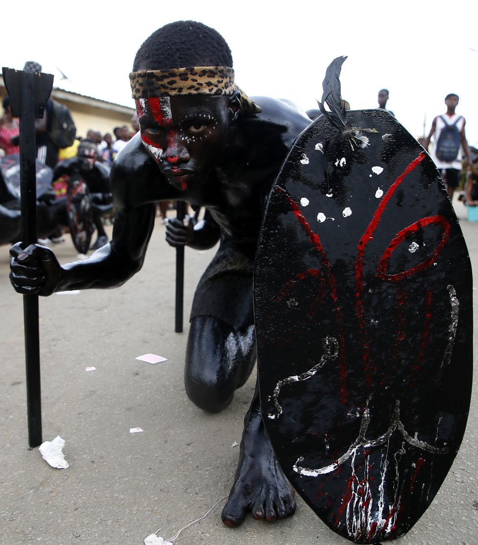 A man painted black crouches on the ground, with a black shield and another stick prop.