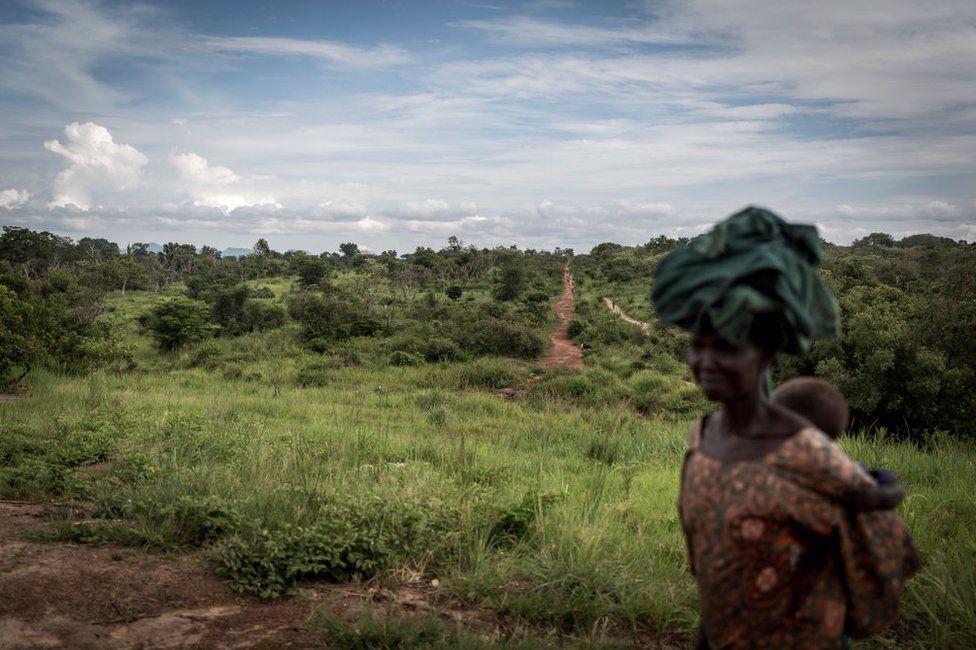 A woman, whose face is blurred, carries an infant on her back as she walks from South Sudan into Uganda.
