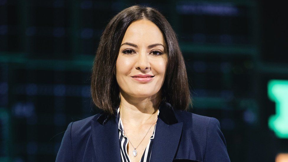 A woman with shoulder length, brown hair wears a navy business suit with a white and blue striped blouse underneath. She's wearing a silver teardrop pendant on a silver chain that rests just below her collarbone. She's smiling, and in the background we can just make out a stage of some sort