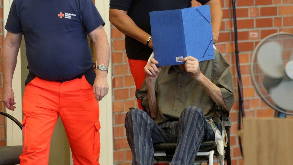 Former Nazi concentration camp guard Josef S (R) hides his face with a folder as he arrives on June 28, 2022 at a gym used as a makeshift courtroom in Brandenburg an der Havel, eastern Germany, where his verdict was spoken.