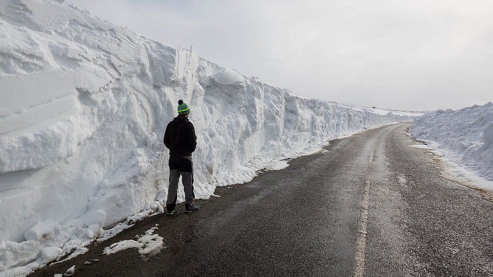 A man stands beneath a huge snow drift on a road in the Pennines on March 20, 2018 in Allenheads, England