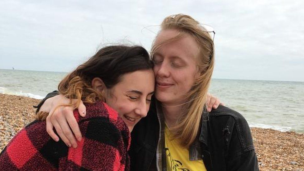 Couple Daisy, 24 and Ramona, 25. Sat on the pebbles of Brighton beach with the sea behind them. They're embracing and both have their eyes closed and are smiling.