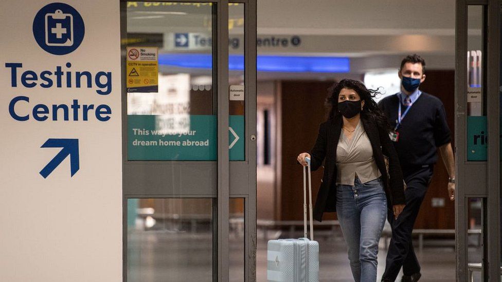 woman with bags at airport wearing mask by testing centre sign