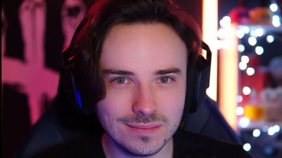 Sammy wears a gaming headset, with various coloured lights of his gaming room visible behind him. He's smiling slightly, and sports a little bit of stubble. His black hair gives off a faint red glow created by the lights in the room around him.