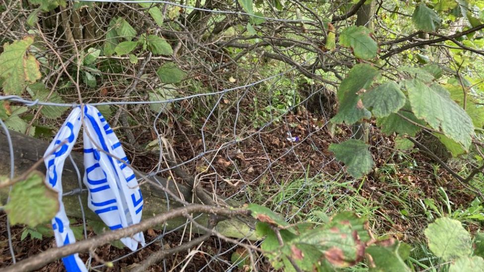White police tape with blue writing on it saying "police" hanging from a metal wire fence in a grassy area near the area of the incident.