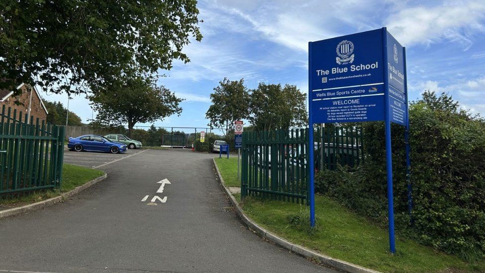 Image of the Blue School in Wells. the road leading into the school can be seen, painted with the word 'in' and an arrow pointing forwards in the direction of travel. There are trees and hedges nearby. A blue metal sign can be seen on the right, printed with 'The Blue School' and the school logo.