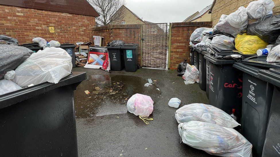 A bin store area on a housing estate showing the wheelie bins full and the bags stacked on top and around