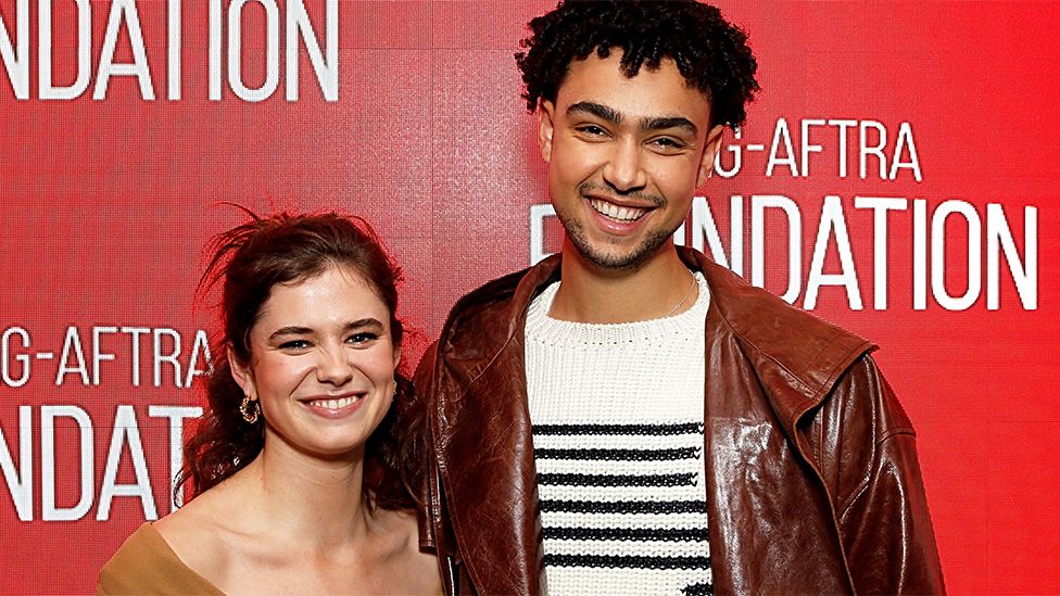 Alison and Archie, standing next to each other with a red background. Alison is smiling, wearing a brown outfit, while Archie, who is taller, is wearing a white and black patterned jumper under a brown leather jacket.
