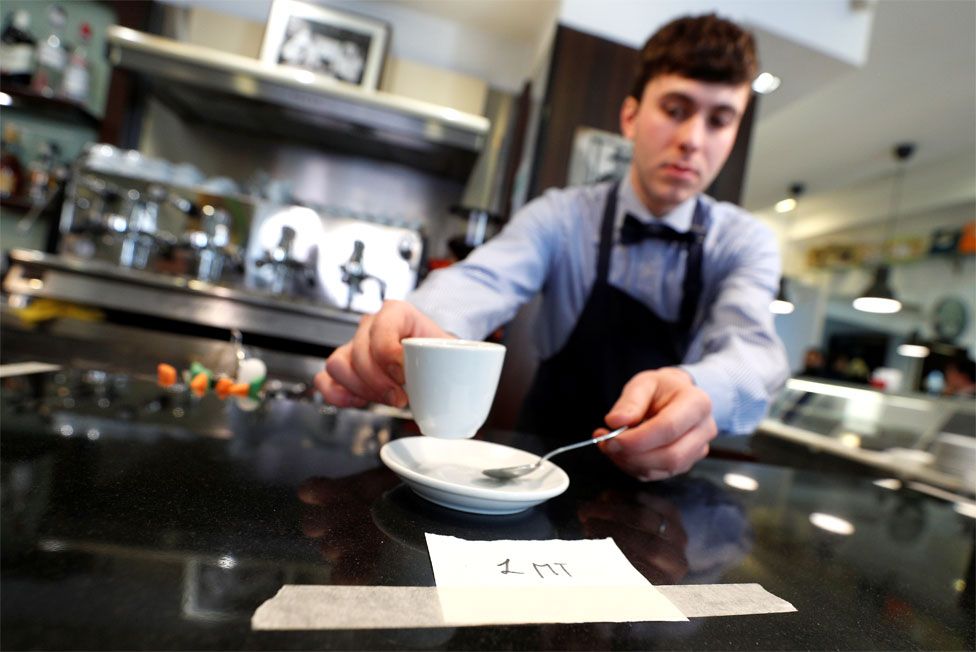 A waiter serves a coffee with a sign saying '1 metre'