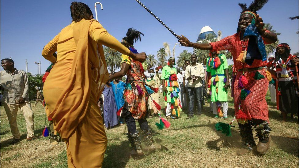 Lively photo of tribal dancers wearing colourful clothing. There is a man in a traditional red outfit with a head dress. He is dancing with a stick and his arms are open wide. A woman opposite him is wearing a yellow outfit and is also dancing. There is a crowd dressed in colourful clothing behind him.