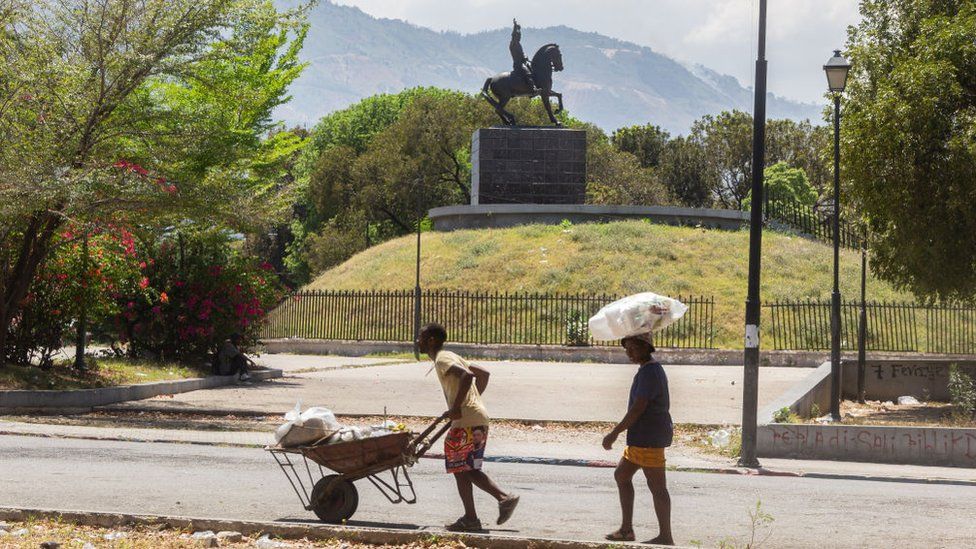 A man passes by with a wheelbarrow of food and a woman behind him carries plastic plates to put food on in the Champs de Mars district, near the National Palace, the city center of port-au-prince is transformed into a veritable battlefield between the forces of law and order and armed gangs.