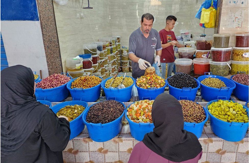 Man serving two female customers at the market. There is an array of colourful food in front of him, stored in blue tubs.