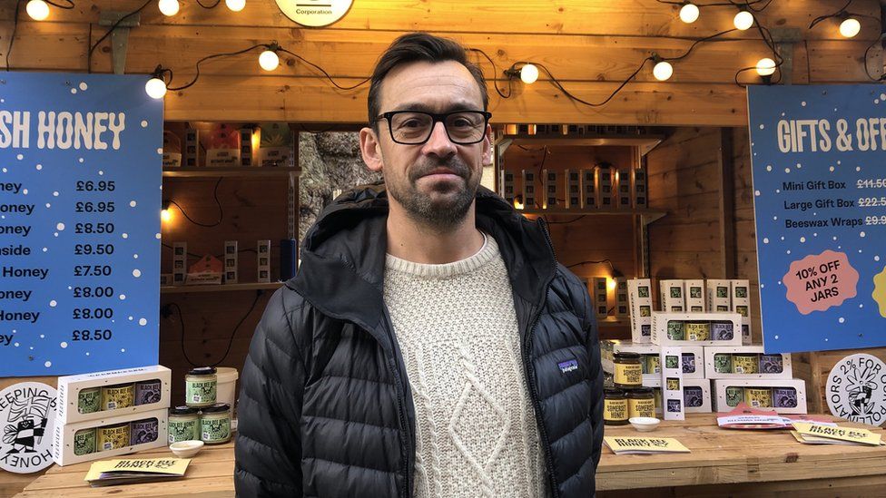 Man in cream sweater and black jacket smiling at camera outside a wooden stall