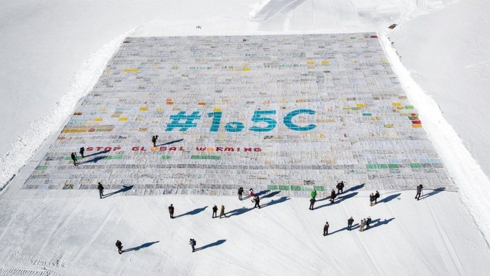 The postcard, seen from above on the snow, reads: "#1.5C - Stop global warming". People walking underneath it look tiny by comparison, emphasising the scale.