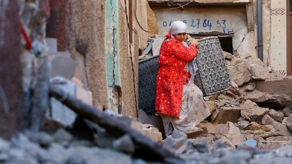 A woman carries furniture through rubble