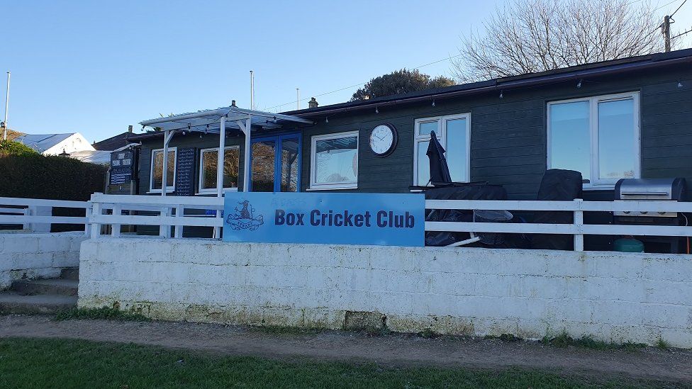 A one storey wooden building with a sign for Box Cricket Club and a blue sky