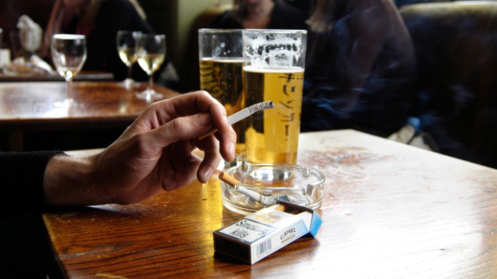 Hand holding a half-smoked cigarette at a pub table, beer glasses in the background