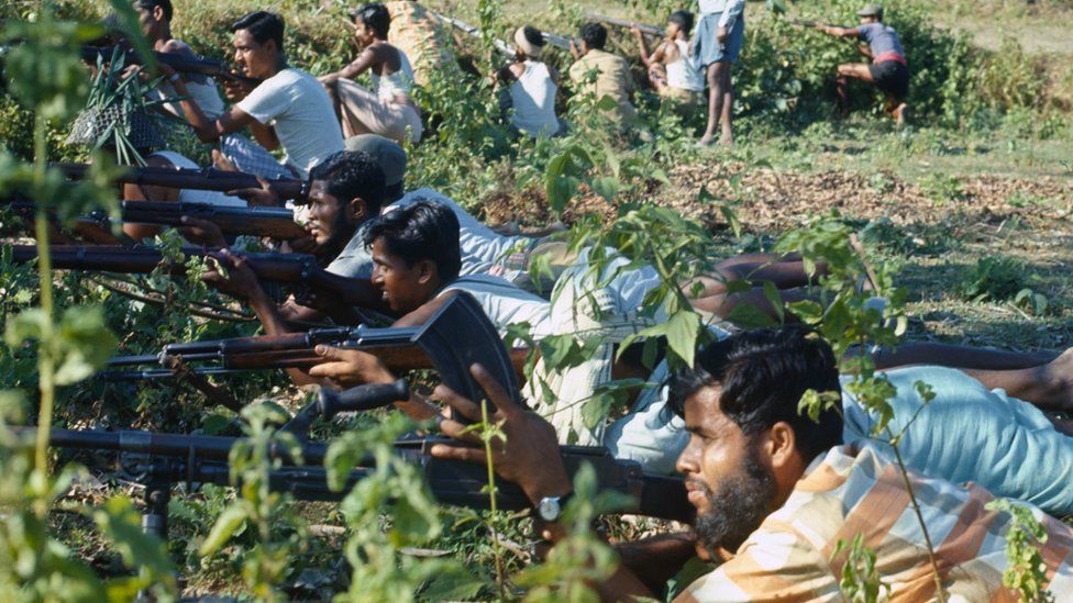 Many Bangladeshi men lined up next to each other, lying on the ground holding rifles