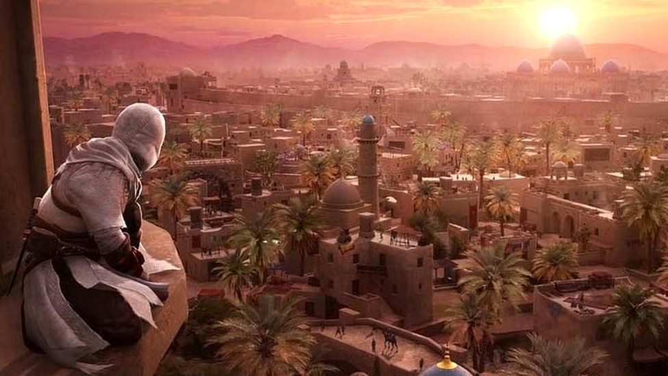 A scene from Assassin's Creed Mirage shows a figure dressed in white, his head covered with a hood, perched on a high ledge on a tall building. He's looking out over 9th Century Baghdad - a sprawling town in the desert. The sun creates a purple glow over the city as it sets, lighting up the collection of flat-topped buildings and domed structures that were typical of the period. The image suggests a vast city to explore