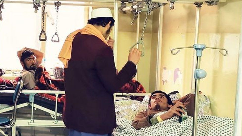 Two injured men in beds in a hospital with one man standing over them holding a chain