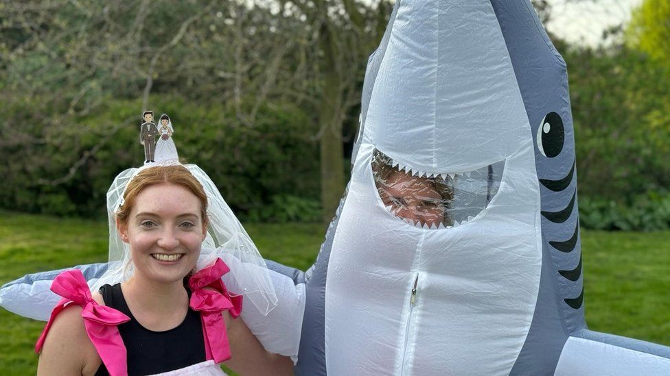 Laura Baker and Georgie Box in their costumes for the London Marathon. Laura is a 27-year-old white woman with strawberry blonde hair which is tied back. She wears a bridal veil and has a wedding cake topper on her head. She has a black sleeveless running top with bright pink straps holding up her costume. Georgie, right, is a 28-year-old white woman and is pictured inside an inflated shark costume with her right arm around Laura. The shark's smiling mouth provides a window through which you can just about make out Georgie's blonde hair. The women are pictured outside in a park on a bright day in front of greenery.