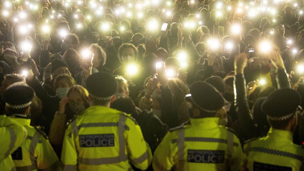 Police stand in front of a crowd of people who turn on their phone torches as they gather in Clapham Common, south London