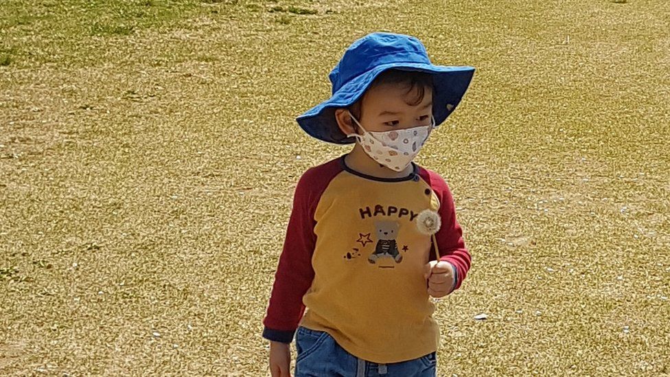 Small boy wearing floppy blue hat and face mask walking on grass