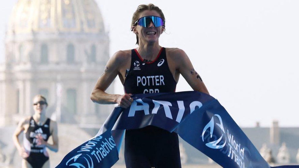 Team GB triathlete Beth Potter crosses the finish line to win the elite women triathlon test event at a test event for the Paris 2024 Olympics. She is wearing a navy Team GB all-in-one suit that goes to just above her kness with "Potter GBR" on the front and has big reflective sunglasses on. Her hair is tied back and she is grimacing slightly as she crosses the blue and white finish line which is a large piece of tape that she's running through.